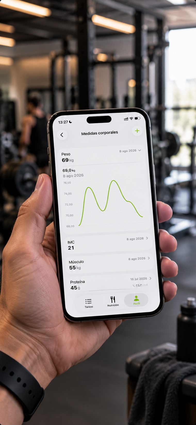 Móvil sostenido en la mano dentro de un gimnasio, mostrando la pantalla de medidas corporales de la app con la gráfica de peso, el IMC y el músculo registrados.